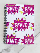 Load image into Gallery viewer, A spiral notebook is on a tabletop. The white spiral notebook has black spiral binding. The pattern design features the word &quot;brave&quot; inside a hot pink star with sketchy elements around. 