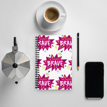 Load image into Gallery viewer, A spiral notebook on a tabletop with a coffee mug and phone around it. The white spiral notebook has black spiral binding. The pattern design features the word &quot;brave&quot; inside a hot pink star with sketchy elements around. 