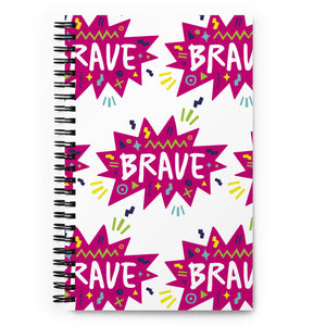 A spiral notebook is on a tabletop. The white spiral notebook has black spiral binding. The pattern design features the word "brave" inside a hot pink star with sketchy elements around. 