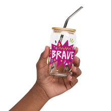 Load image into Gallery viewer, A photo of a glass can tumbler being held in a hand. The glass tumbler has a bamboo lid and silver metal straw. The design on the glass tumbler says the word &quot;brave&quot; inside a hot pink star with illustrated sketchy elements around the word. 