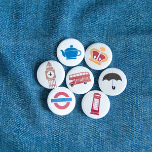 A set of seven buttons on a jean jacket. The buttons features illustrations of a red telephone booth, Big Ben, a teapot, a royal crown, a double decker bus, a black umbrella and the London underground/tube logo. 