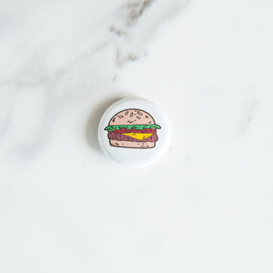 A button pin on a white tabletop. The pin features an illustration of a hamburger.