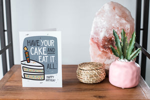 A card on a wood tabletop and on the right side of the card is a woven basket, a pink plant pot with a cactus in it and a pink crystal rock. The card features the words "Have your cake and eat it all, happy birthday” with an illustrated piece of cake with a candle on the top.