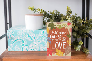 A greeting card is on a table top with a present in the background. There's greenery on top of the present. The card reads "May Your Gathering Be Filled with Love" with the words inside an illustrated pumpkin with leaves surrounding the pumpkin.