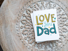 Load image into Gallery viewer, A greeting card laying on a wooden table with some cut wood details. The card features the words “Love you Dad” with small stars around the letters. 