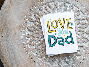 A greeting card laying on a wooden table with some cut wood details. The card features the words “Love you Dad” with small stars around the letters. 