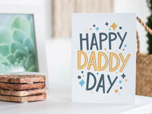 Load image into Gallery viewer, A greeting card featured standing up on a white tabletop with a framed photo of a succulent in the background and a stack of wooden coasters. There’s a woven basket in the background with a cactus inside. The card features the words “Happy Daddy Day” with diamond shapes surrounding the letters. 