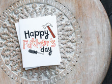 Load image into Gallery viewer, A greeting card laying on a wooden table with some cut wood details. The card features the words “Happy Father’s Day” with an illustrated hammer and screwdriver around the words. 
