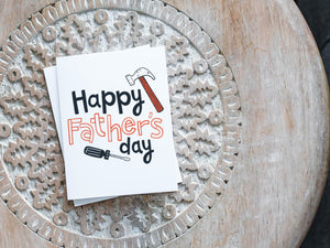 A greeting card laying on a wooden table with some cut wood details. The card features the words “Happy Father’s Day” with an illustrated hammer and screwdriver around the words. 