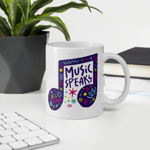 A white mug on a desktop. The mug features a design with the words 'music speaks' in an illustrated musical note with sketchy elements around it. 