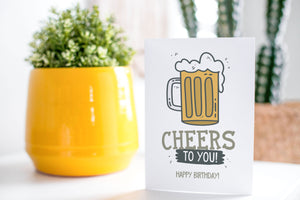 A greeting card is on a table top with a yellow plant pot and a green plant inside. The card features the words “Cheers to you, happy birthday” with an illustrated beer mug.