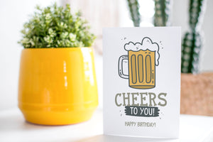A greeting card is on a table top with a yellow plant pot and a green plant inside. The card features the words “Cheers to You! Happy Birthday!” with an illustrated beer mug.