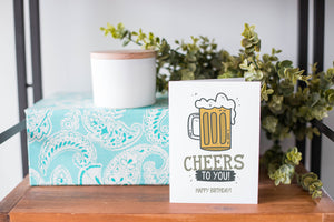 A greeting card is on a table top with a present in blue wrapping paper in the background. On top of the present is a candle and some greenery from a plant too. The card features the words “Cheers to You! Happy Birthday!” with an illustrated beer mug.