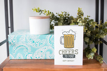 Load image into Gallery viewer, A greeting card is on a table top with a present in blue wrapping paper in the background. On top of the present is a candle and some greenery from a plant too. The card features the words “Cheers to you, happy birthday” with an illustrated beer mug.