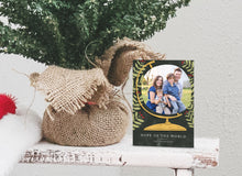 Load image into Gallery viewer, A photo of a one-sided Christmas card showing the front of the card standing up next to the bottom of a small Christmas tree with the base wrapped in burlap. The photo card features a photo inside an illustrated globe. Around the globe are illustrated leaves. At the bottom of the globe the words “Hope of the World.”