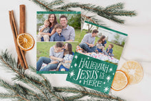 Load image into Gallery viewer, A photo of a double-sided Christmas card showing the front and back of the card laying on a white surface. Around the two sides of the card are pine needles, cinnamon sticks and dried oranges. The front of the card features a photo on the top and on the bottom is in green with words in white “Hallelujah Jesus is Here” with white stars around it. The back of the card features two photos. 