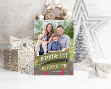Load image into Gallery viewer, A photo of a one-sided Christmas card showing the front of the card standing up with Christmas items behind the card. There’s a sparkly star, glitter wrapped gift, ribbon and ornaments with a small silver tree around the photo card. The photo card features one photo with the words “O Come Let Us Adore Him” below with illustrated red leaves.