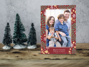 A photo of a one-sided Christmas card showing the front of the card standing up with three small Christmas trees next to it. The photo card features one photo with a frame of illustrated leaves in various colors. Below the photo it reads “Rejoice Greatly” and has space to put your family name. 