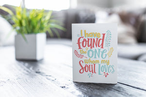 A greeting card featured on a black, wood coffee table. There’s a white planter in the background with a green plant. There’s also a gray sofa in the background with a white pillow. The card features the words “I have found the one whom my soul loves.”