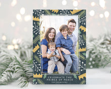 Load image into Gallery viewer, A photo of a one-sided Christmas card showing the front of the card standing up with pine needles behind and blurred white Christmas lights. The photo card features one photo with a border of illustrated gold crowns, and green and white illustrated leaves. The bottom of the card reads “Celebrate the Prince of Peace, The Thompsons.”
