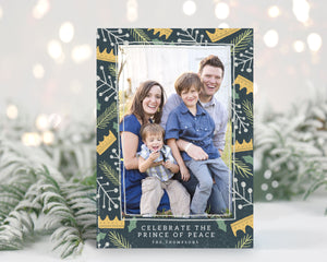 A photo of a one-sided Christmas card showing the front of the card standing up with pine needles behind and blurred white Christmas lights. The photo card features one photo with a border of illustrated gold crowns, and green and white illustrated leaves. The bottom of the card reads “Celebrate the Prince of Peace, The Thompsons.”