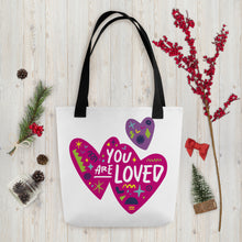 Load image into Gallery viewer, A white tote bag with black handles on a wood tabletop with Christmas items around it. The bag features a design with the words &#39;you are loved&#39; inside two illustrated pink hearts with a smaller, purple heart above. 