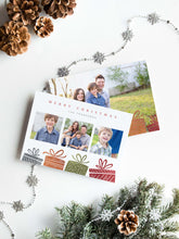 Load image into Gallery viewer, A photo of a double-sided Christmas card showing the front and back of the card laying on a white surface. Around the two sides of the card are pine cones, pine needles and a string of silver snowflake garland. The front of the card features three photos with illustrated gifts on the bottom. On top of the photos the words “Merry Christmas” is featured with a family name below which you can edit. The back of the card features one photo with some more illustrated gifts. 