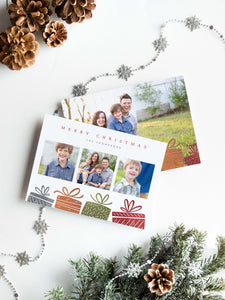 A photo of a double-sided Christmas card showing the front and back of the card laying on a white surface. Around the two sides of the card are pine cones, pine needles and a string of silver snowflake garland. The front of the card features three photos with illustrated gifts on the bottom. On top of the photos the words “Merry Christmas” is featured with a family name below which you can edit. The back of the card features one photo with some more illustrated gifts. 