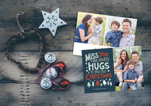 A photo of a Christmas card showing the front and back of the card laying on a wood surface. To the left of the cards are some ornaments. The front of the card features a photo on the right side and on the left side are the words “ Miss you and sending hugs to you this Christmas” with illustrated trees below the words. The back of the card features two photos with illustrated trees at the bottom.