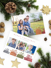 Load image into Gallery viewer, A photo of a two-sided Christmas card showing the front of the card on top of a brown wrapped gift on a white tabletop. Around the gift are pine needles, pinecones and wood star ornaments. The front of the card features three photos with illustrated gifts on the bottom. On top of the photos the words “Merry Christmas” is featured with a family name below which you can edit. The back of the card features one photo with some more illustrated gifts.