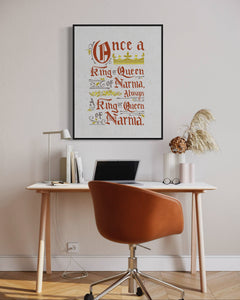 Artwork in a black frame with the with a white matte. The frame is on a wall above a desk. The artwork features hand drawn lettering of the Narnia quote "Once a king or queen of Narnia, always a king or queen of Narnia."