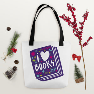 A white tote bag with black handles on a wood tabletop with Christmas items around it. The tote bag has a design with the words 'I love books' with the word love shown as a heart. The words are inside an illustrated book. 