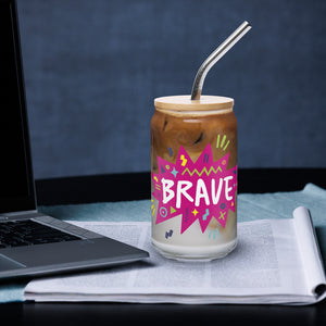 A photo of a glass can tumbler on a desktop next to a laptop. The glass tumbler has a bamboo lid and silver metal straw. The design on the glass tumbler says the word "brave" inside a hot pink star with illustrated sketchy elements around the word. 