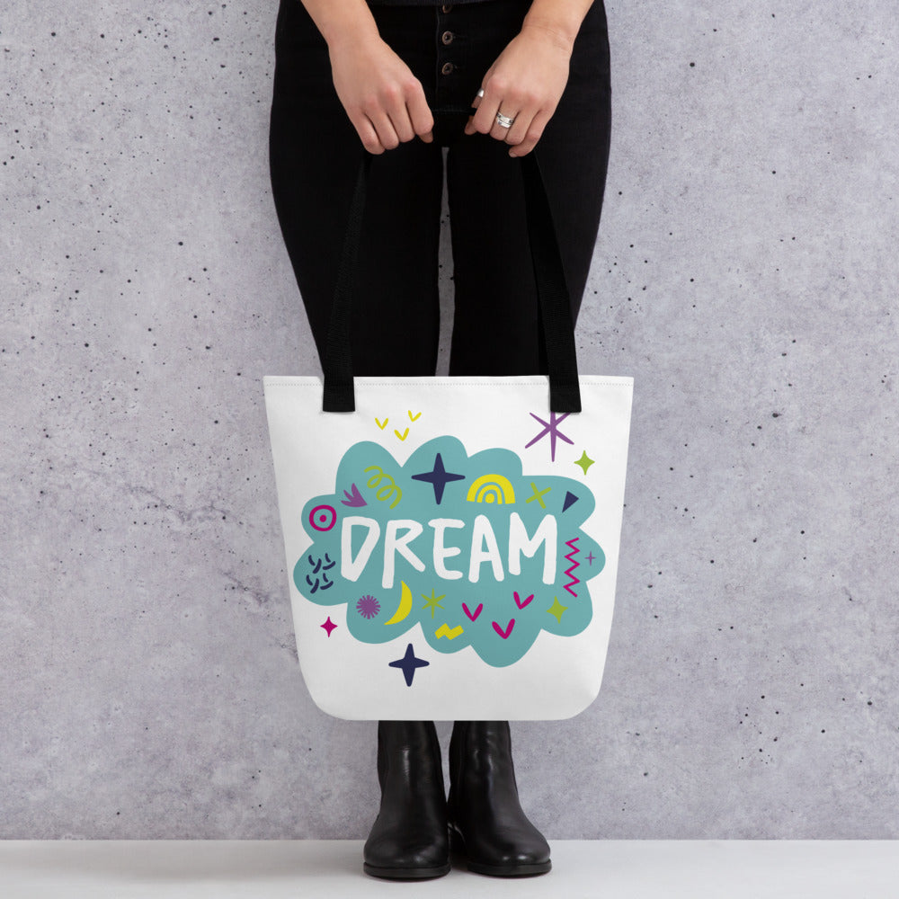 A person holding a white tote bag with black handles. The bag features a design with the word 'dream' with an illustrated cloud around the word with colorful sketchy around. 