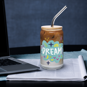 A photo of a glass can tumbler on a desktop next to a laptop. The glass tumbler has a bamboo lid and silver metal straw. The design on the glass tumbler says the word "dream" inside a teal cloud illustration with sketchy illustrations around. 