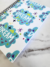 Load image into Gallery viewer, A spiral notebook on a tabletop. The white spiral notebook has black spiral binding. The pattern design features the word &quot;dream&quot; inside a cloud shape with sketchy elements around. 