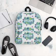 Load image into Gallery viewer, A backpack on a table with a pattern design with the word &quot;dream&quot; inside a cloud illustration with sketchy elements. The bag is surrounded by a laptop, headphones, shoes, a phone and a notebook. 