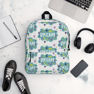 A backpack on a table with a pattern design with the word "dream" inside a cloud illustration with sketchy elements. The bag is surrounded by a laptop, headphones, shoes, a phone and a notebook. 