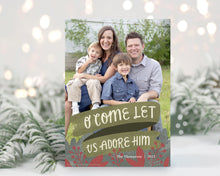 Load image into Gallery viewer, A photo of a one-sided Christmas card showing the front of the card standing up with pine needles behind and blurred white Christmas lights. The photo card features one photo with the words “O Come Let Us Adore Him” below with illustrated red leaves. 