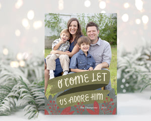 A photo of a one-sided Christmas card showing the front of the card standing up with pine needles behind and blurred white Christmas lights. The photo card features one photo with the words “O Come Let Us Adore Him” below with illustrated red leaves. 