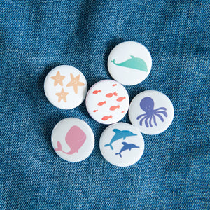 A set of six buttons on a jean jacket. The buttons feature illustrations of whales, fish, starish, dolphins, and an octopus. 