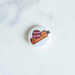 A buttons on a white tabletop. The button features an illustrations of a cricket ball and bat. 