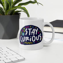 Load image into Gallery viewer, A white mug on a desktop. The mug features a design with the words &#39;stay curious&#39; in an illustrated oval shape with sketchy elements around it. 