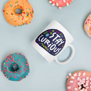 A white mug laying on a tabletop with doughnuts around it. The mug features a design with the words 'stay curious' in an illustrated oval shape with sketchy elements around it. 