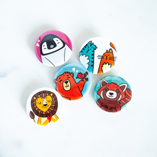 Five button pins are on a white tabletop. The buttons feature illustrated cats, a bear, a penguin, a lion and a red panda.