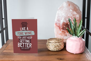 A greeting card featured standing up on a white tabletop with a pink plant pot in the background and some succulents in the pot. There’s a woven basket in the background with a cactus inside. The card features the words “Like a Fine Wine, You Just Keep Getting Better, Happy Birthday.”