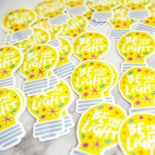 Load image into Gallery viewer, Die cut vinyl stickers with the words &quot;be the light&quot; inside an illustrated lightbulb.