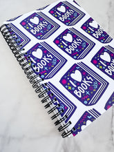 Load image into Gallery viewer, A spiral notebook on a tabletop. The white spiral notebook has black spiral binding. The pattern design features the words &quot;I love books&quot; inside an illustrated book with the word love as a heart. 