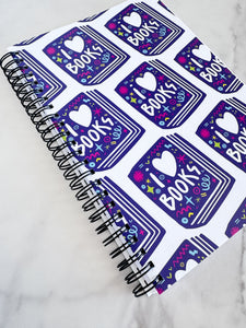 A spiral notebook on a tabletop. The white spiral notebook has black spiral binding. The pattern design features the words "I love books" inside an illustrated book with the word love as a heart. 