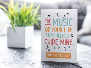 A greeting card is featured on a wood coffee table with a green plant in a white planter in the background. The card features the words “The music of your life has helped guide mine. Happy Father's Day.” 